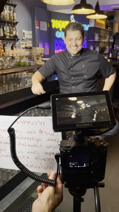 Monitor de cámara enfocando a un entrevistado sonriente detrás de la barra, con notas del guión escritas a mano en primer plano durante un rodaje de vídeo corporativo