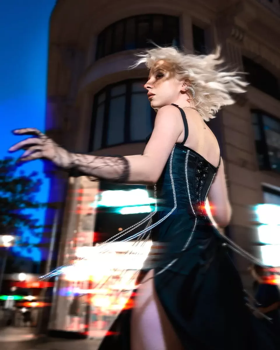 Modelo con vestido negro y guante de encaje girando de noche en una calle de ciudad, foto de cuerpo entero en encuadre vertical con estelas de luz de los coches